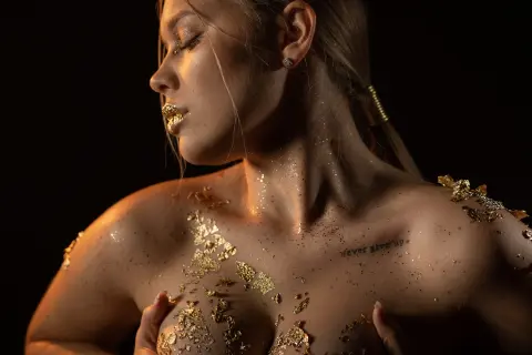 Close-up profile of a woman with gold glitter skin and gold leaf accents against a dark moody background.