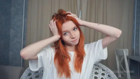 Redheaded woman in a white t-shirt smiling thoughtfully in a bright indoor setting.