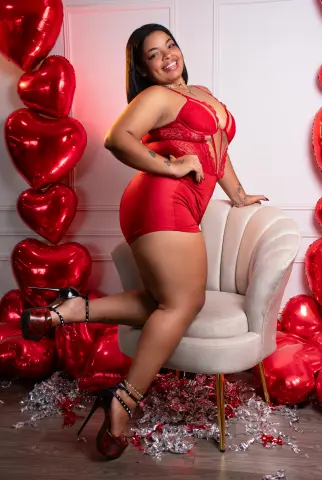 Smiling woman in red bodysuit sitting in a white chair surrounded by red heart balloons for Valentine's Day.