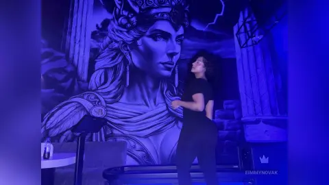 Woman in black workout clothes dancing in front of a large blue goddess mural in a moody, neon-lit studio.