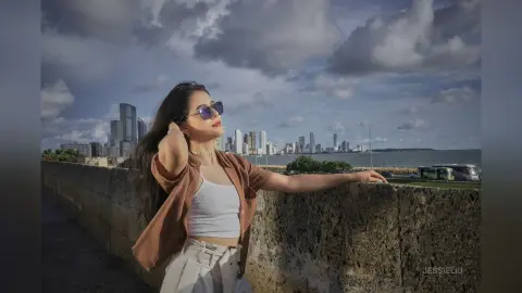 JessieLiu leans against a stone wall outdoors wearing a brown open shirt and a white crop top under a cloudy sky.