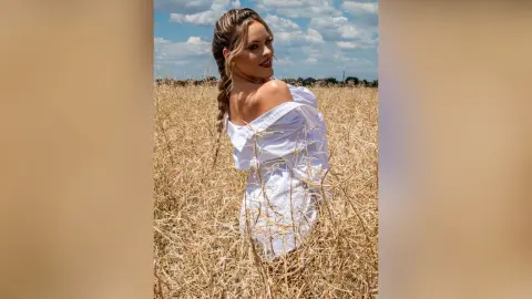Blonde woman in a white off-the-shoulder dress standing in a golden wheat field under a blue sky.