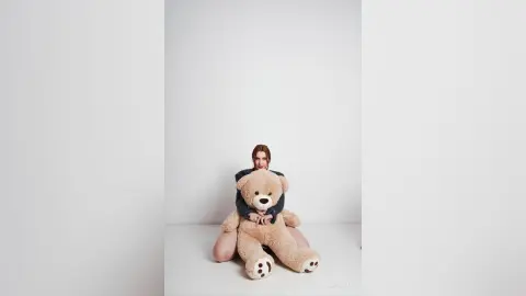 AryaSin sits on the floor against a white background, hugging a large tan teddy bear while wearing a dark grey long-sleeved top.