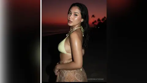 LaurenKaterine poses on a dark beach at sunset, wearing a light yellow bikini top and a patterned skirt, showing off her bare back and shoulders.