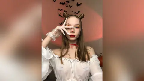 Woman in white gothic dress with red makeup and bat decor for a spooky Halloween aesthetic.