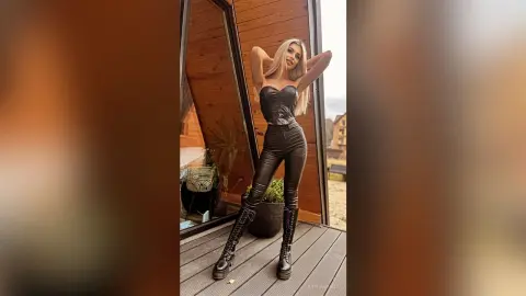Stylish woman in black leather bodysuit and lace-up boots posing confidently on a wooden balcony.