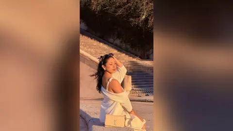 Stylish woman in a white knit sweater and sunglasses sitting on stone stairs during a warm sunset.