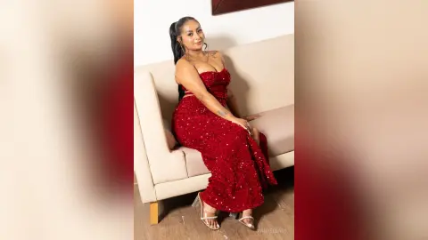 Elegant woman in a sparkling red sequin evening gown posing on a beige sofa in a bright indoor setting.