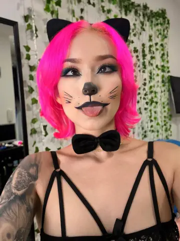 Woman with pink hair and cat face makeup wearing a black lace outfit and ears in a garden setting.