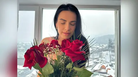Serene woman in pink holding a bouquet of red roses by a window overlooking a snowy landscape.