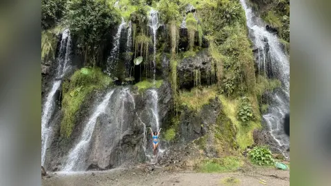 Woman in a blue bikini standing before a lush, green tropical waterfall in a serene natural setting.