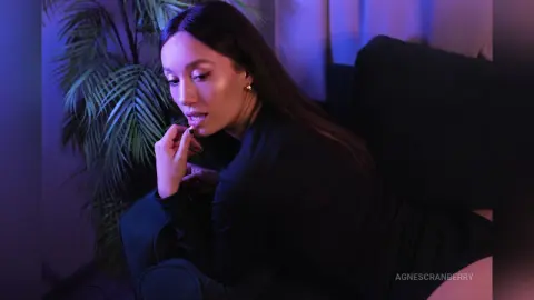 Elegant woman in a black outfit relaxing on a sofa in moody blue lighting near a tropical palm plant.