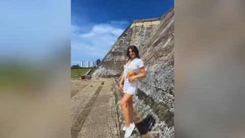 LauraRiveiro — LauraRiveiro stands outdoors in a white crop top and shorts against a large stone wall under a blue sky.