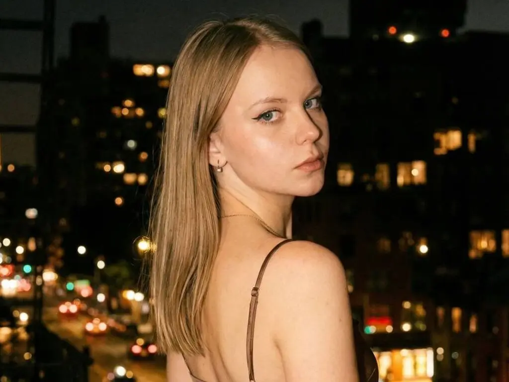 SophiePeachy looks over her shoulder with long blonde hair against a dark city skyline at night.