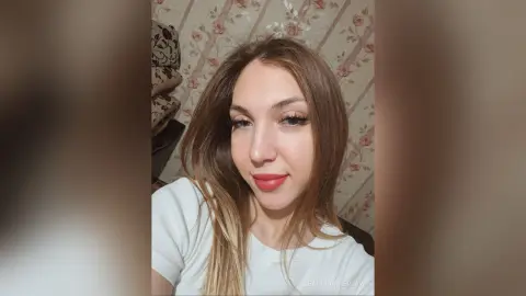 Young woman with long blonde hair wearing a white t-shirt in a room with floral wallpaper.