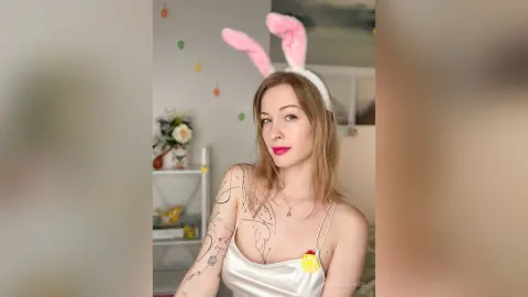 Smiling woman in a white satin camisole wearing pink bunny ears in a bright, cozy indoor setting.
