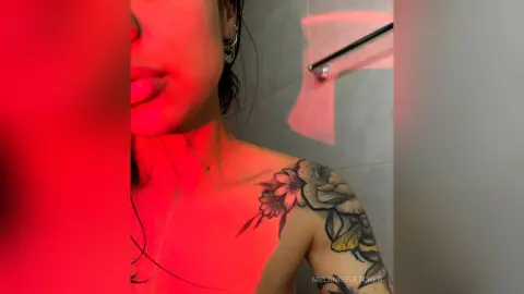 Young woman with shoulder tattoos posing under moody red lighting in a modern bathroom setting.