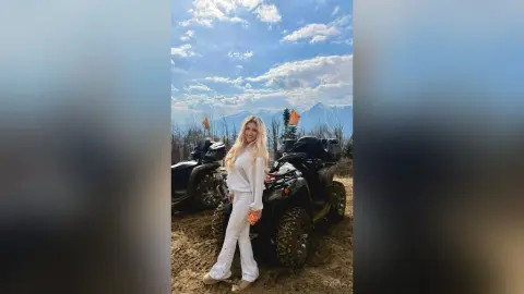 Anita stands outdoors in a white long-sleeve top and white pants next to a black ATV under a blue sky.