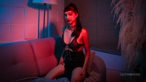 LilithBrooks poses on a light-colored sofa wearing a black lace bra and a black corset-style skirt or bodysuit.