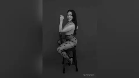 Smiling woman in patterned leggings sitting on a stool in a moody black and white studio portrait.