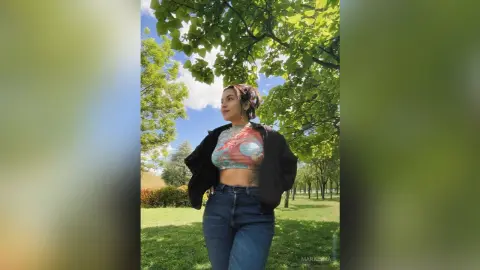 Smiling young woman in a colorful crop top and blue jeans standing under lush green trees in a sunny park.