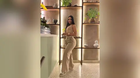 Elegant woman in beige jumpsuit standing in a modern minimalist interior with indoor plants.