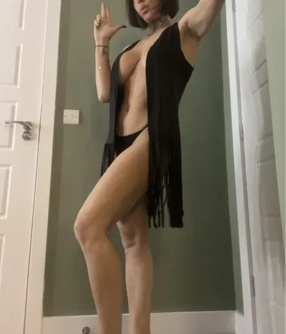 Dee_Licious — Dee_Licious stands in a black fringed dress that is open to show her bare breasts and stomach.