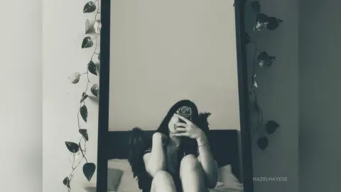 Moody black and white mirror selfie of a woman in a dark outfit sitting in a minimalist bedroom with vine decor.