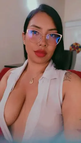 Young woman with glasses and a white sleeveless shirt posing indoors with a confident mood.