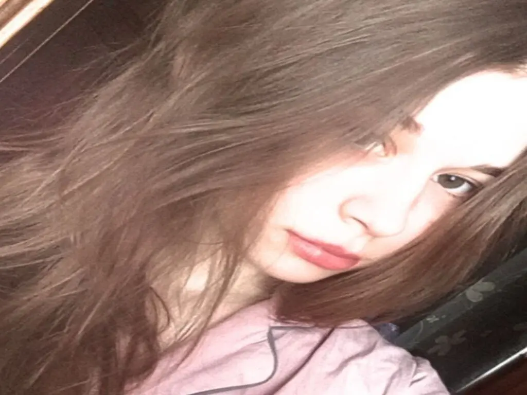 Young woman with long brown hair wearing a pink shirt in a soft, moody indoor setting.