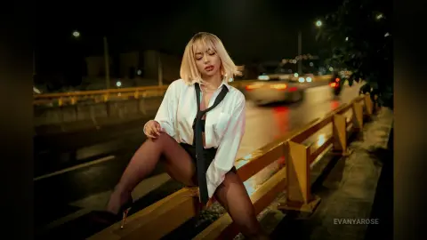 Blonde woman in a white button-down shirt and black shorts sitting on a city street railing at night.