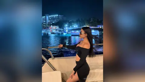 Stylish woman in a black cutout dress posing on a boat at night with city lights reflecting on the water.