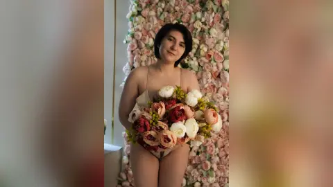 Young woman in beige lingerie holding a large bouquet of roses against a romantic floral wall background.
