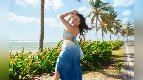 Smiling woman in a blue bikini and sarong walking through tropical palm trees on a sunny beach path.