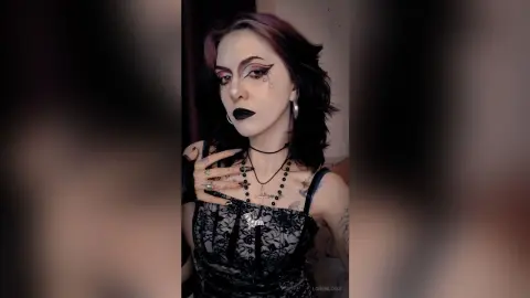 Woman with goth makeup and dark wavy hair wearing a black lace camisole and silver necklaces in a moody setting.