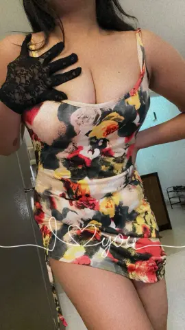 Darling_Mona wears a floral print mini dress and black lace gloves while posing in a room.