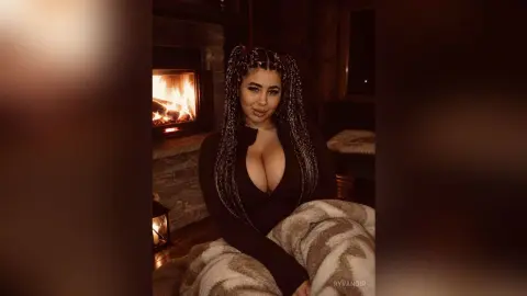 Woman with long braids in a cozy dark outfit relaxing by a warm glowing fireplace.