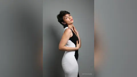 IveteDurand poses in a tight black and white sleeveless dress, looking over her shoulder with her hands resting on her chest and neck.