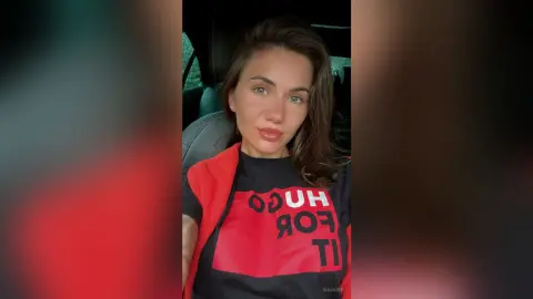 Young woman in a black Hugo Boss graphic t-shirt and red vest sitting in a car with a relaxed expression.