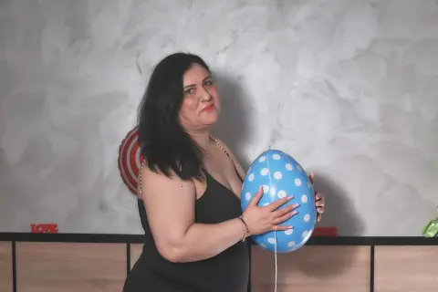 Smiling woman in a black dress holding a blue polka dot balloon against a grey textured wall.