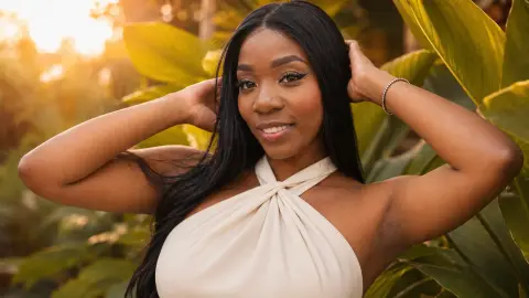 Smiling Black woman in a white halter top posing amidst lush tropical greenery at sunset.