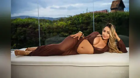 Glamorous blonde woman in a brown sheer maxi dress relaxing on a white lounge chair outdoors at sunset.