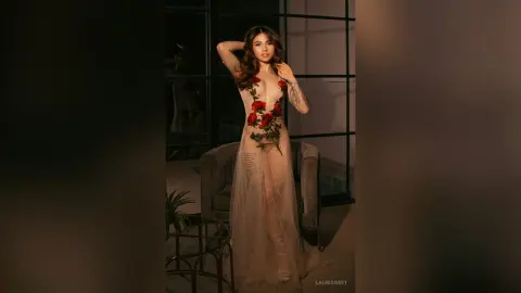 Elegant woman in a sheer floral gown posing in a moody, dimly lit modern interior.