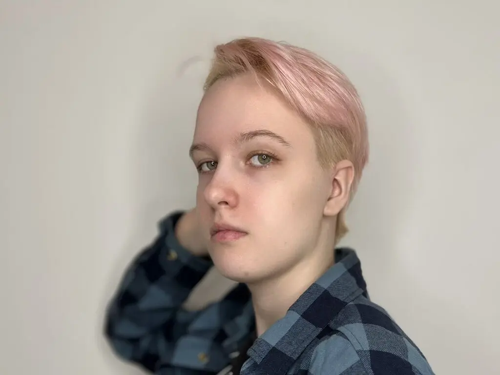 Young woman with short pink hair wearing a blue plaid shirt against a neutral white background.