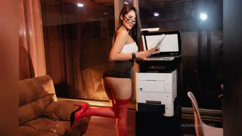 VeronicaValdez stands by a white printer wearing a black tube top and black skirt with red thigh-high stockings.