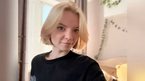 Blonde woman in a black t-shirt smiling in a bright, sunlit room with indoor plants.