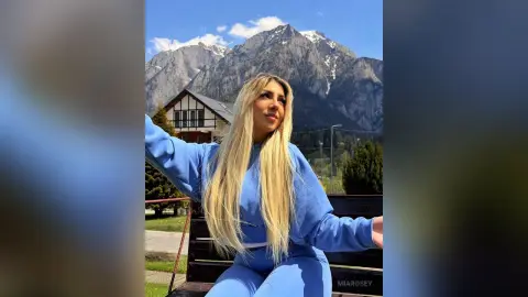 Blonde woman in a blue sweatshirt relaxing outdoors against a scenic mountain landscape on a sunny day.