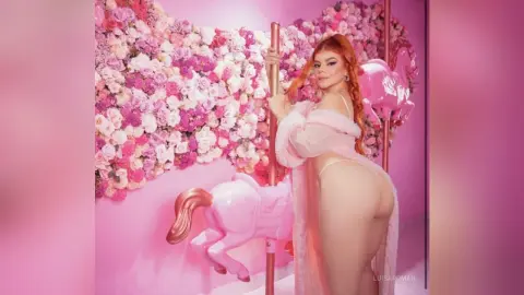 Glamorous woman in a pink fuzzy outfit posing by a pink unicorn statue against a floral rose wall.