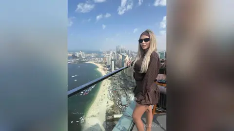 Stylish woman in a brown dress and sunglasses enjoying a sunny view from a high-rise balcony overlooking a coastal city.