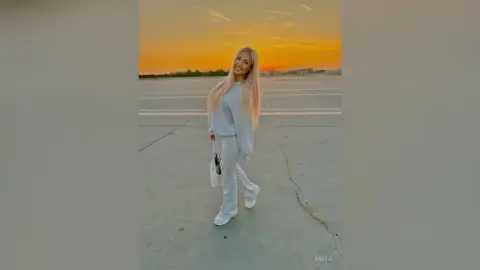 Anita stands on a paved road at sunset wearing a light grey long-sleeve top and matching trousers.